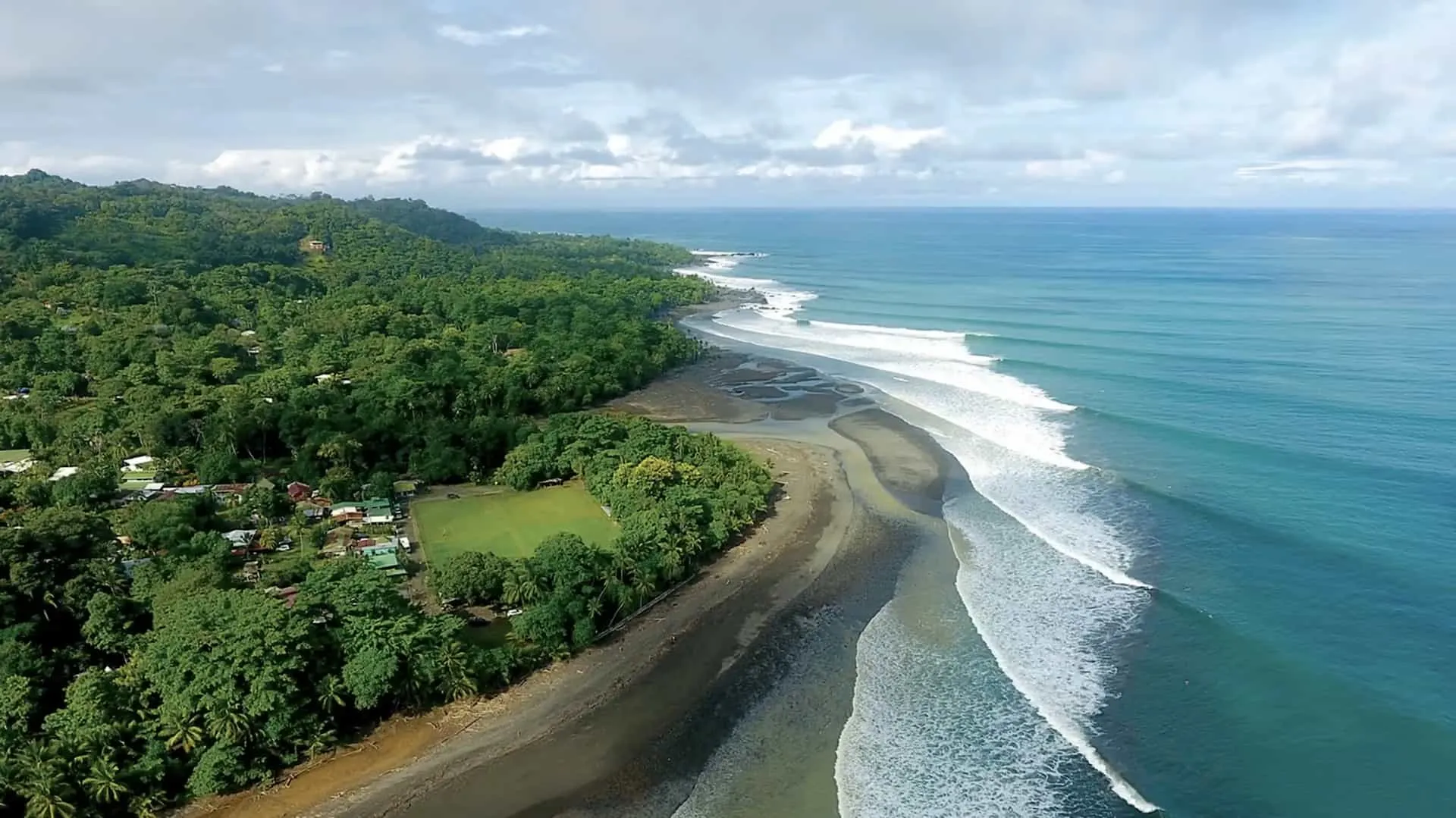 Wide view of the Pavones left-hand point break with a surfer mid-ride, jungle coastline in the background